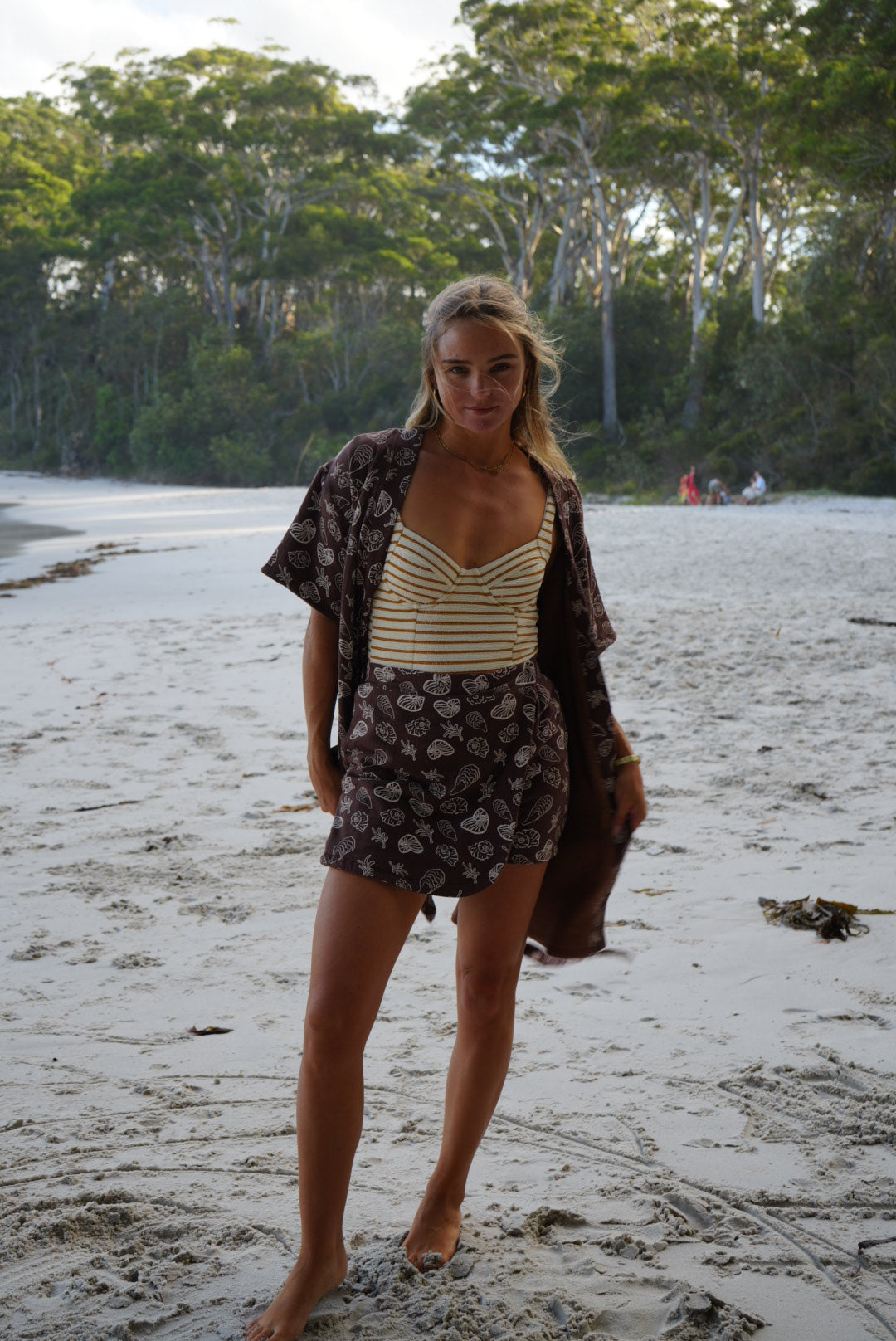 Woman standing on a sandy beach with trees in the background wearing striped swimwear, brown with seashell design matching Towel Skort and Towel Robe