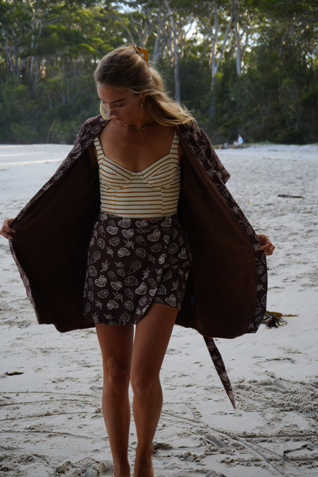 Woman standing at beach in Brown Towel Skort and Brown Seashell Towel Robe with striped swimwear on 